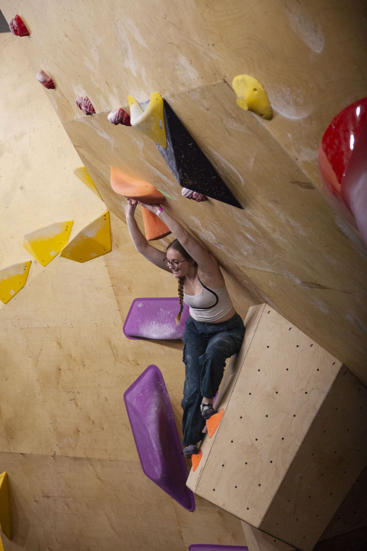 This image is a photograph of a female climber from a previous climbing competition at Highball Climbing Centre. It shows the climber with both hands touching a hold above her head. She is facing away from the wall, smiling, presumably at other climbers waiting to go on the wall. This image is used as an example of routes set for competitions and shows a climber enjoying herself at an event.