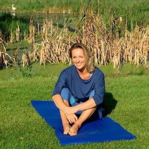 This image is of Avril. She is wearing a loose blue top and light blue yoga leggings. She is sat outside, with reeds behind her. She is sat on a dark blue yoga mat, which is laying on the grass. She is smiling at the camera, with the sun in her eyes.