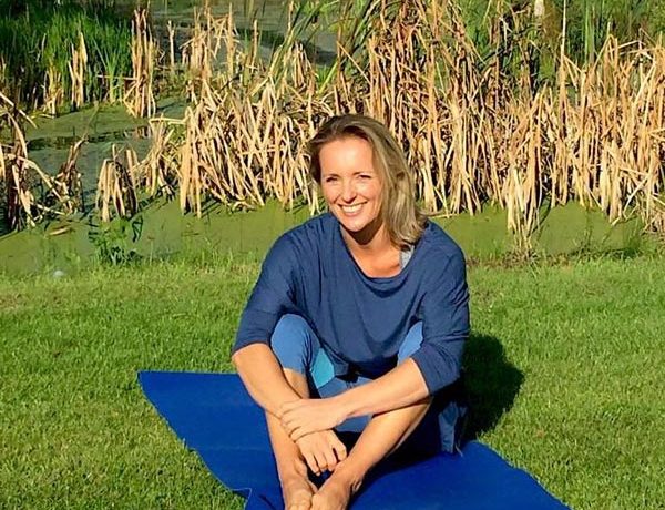 This image is of Avril. She is wearing a loose blue top and light blue yoga leggings. She is sat outside, with reeds behind her. She is sat on a dark blue yoga mat, which is laying on the grass. She is smiling at the camera, with the sun in her eyes.