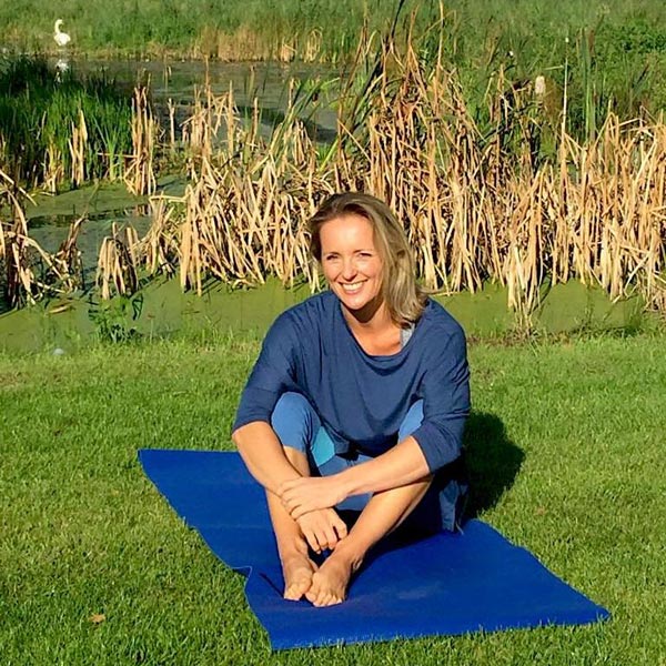 This image is of Avril. She is wearing a loose blue top and light blue yoga leggings. She is sat outside, with reeds behind her. She is sat on a dark blue yoga mat, which is laying on the grass. She is smiling at the camera, with the sun in her eyes.
