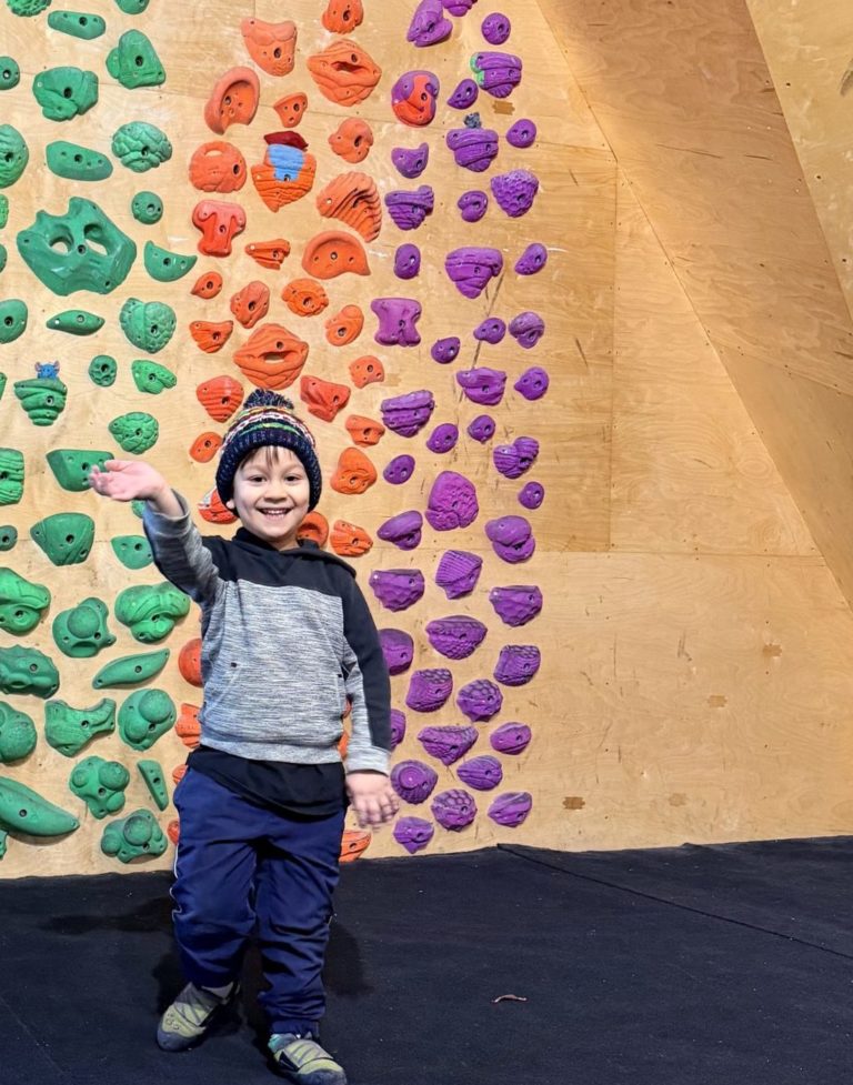 This is an image of a child standing in front of a short climbing wall. The child is waving at the camera and smiling.